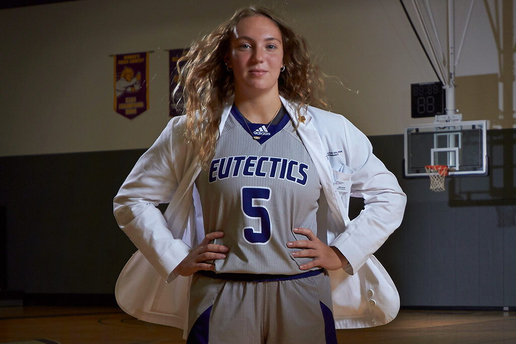 UHSP student Grace Beyer poses in her basketball uniform and pharmacy white coat in the UHSP competition gym